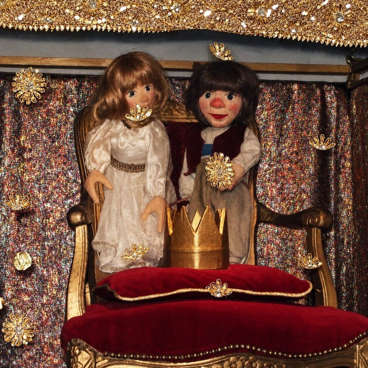 Zwei Puppen in glitzernder Kulisse vor einem roten Thron